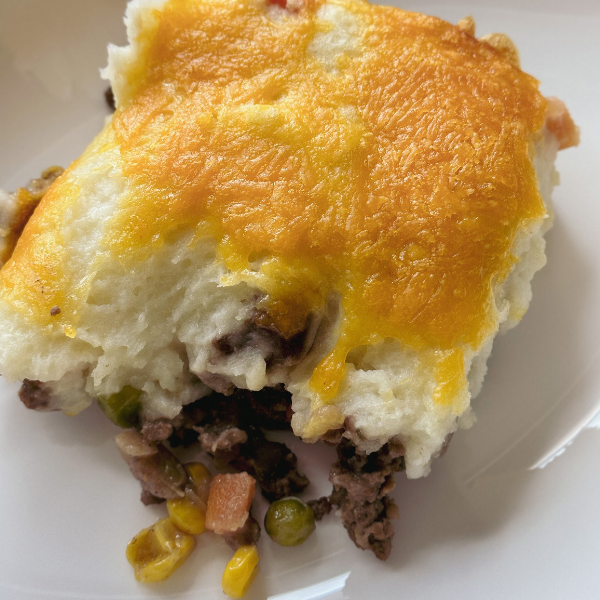 freshly baked shepherd's pie on a white plate golden mashed potato crust on top is slightly crispy revealing layers of ground meat vibrant green peas yellow corn and bright orange carrots