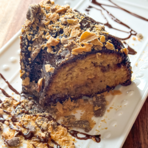 A close-up of a slice of Butterfinger Bundt Cake on a white plate. The cake is moist and golden, topped with a glossy chocolate frosting and sprinkled with crushed Butterfinger candy pieces. Chocolate drizzle decorates the plate, adding an elegant touch to the presentation. The texture of the cake and the crunchiness of the candy create a visually appealing dessert.