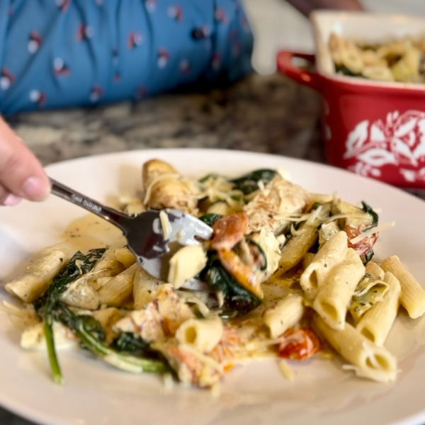 A plate of creamy Tuscan chicken pasta with penne, spinach, and cherry tomatoes. A fork is lifting a bite of pasta and spinach. In the background, a red casserole dish filled with more pasta."